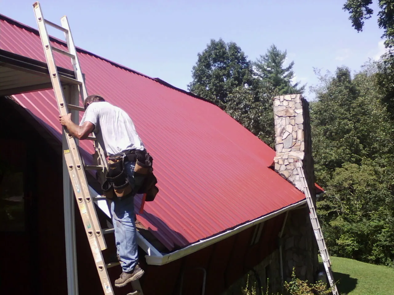 Roofer on a ladder beside a red metal roof and stone chimney — Soffit & Fascia Repair by Roofing Brothers in Seven Springs